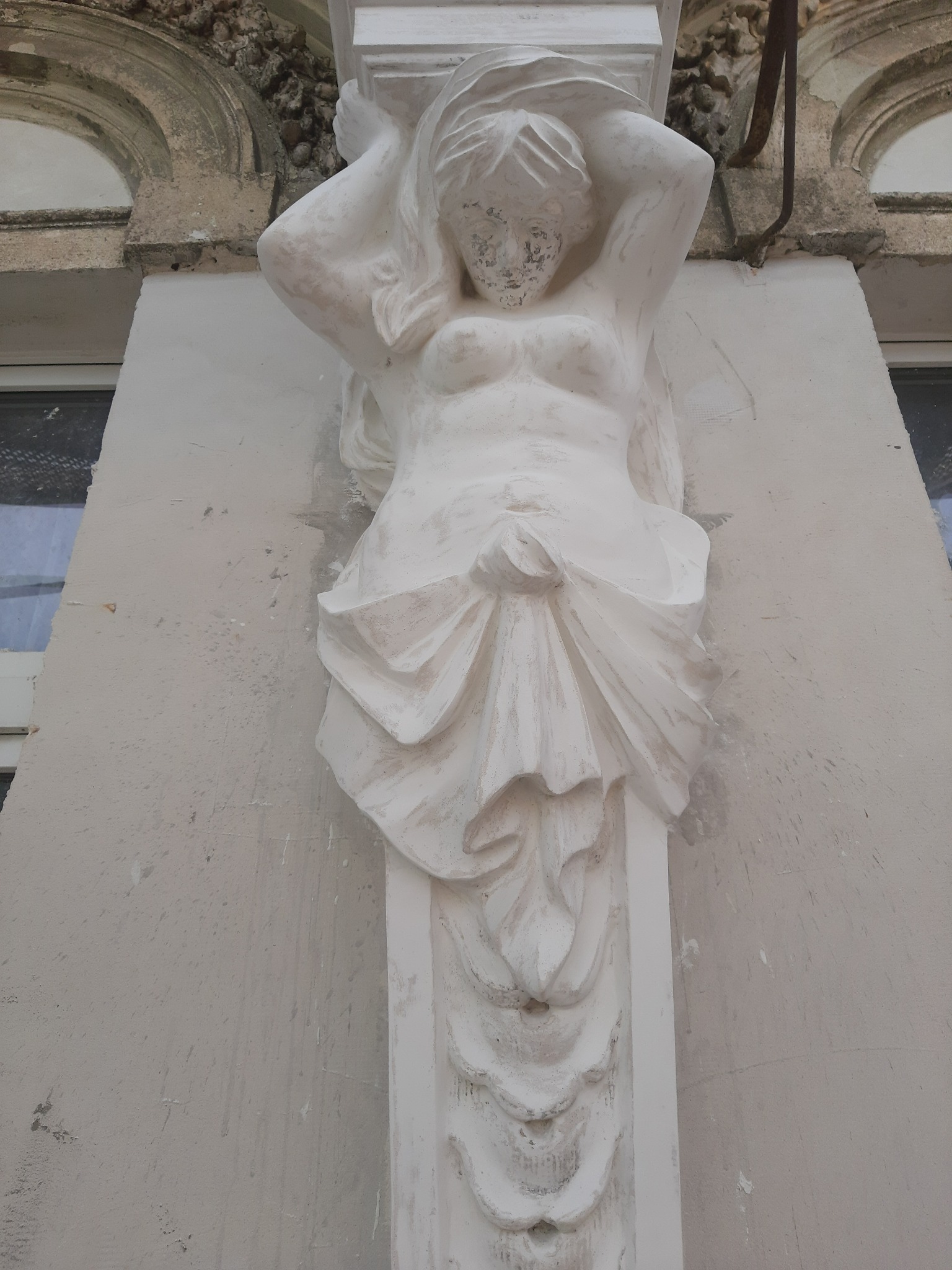 Biała, kobieca figura podtrzymująca element architektoniczny elewacji, widoczne ślady zabrudzeń i odprysków farby.