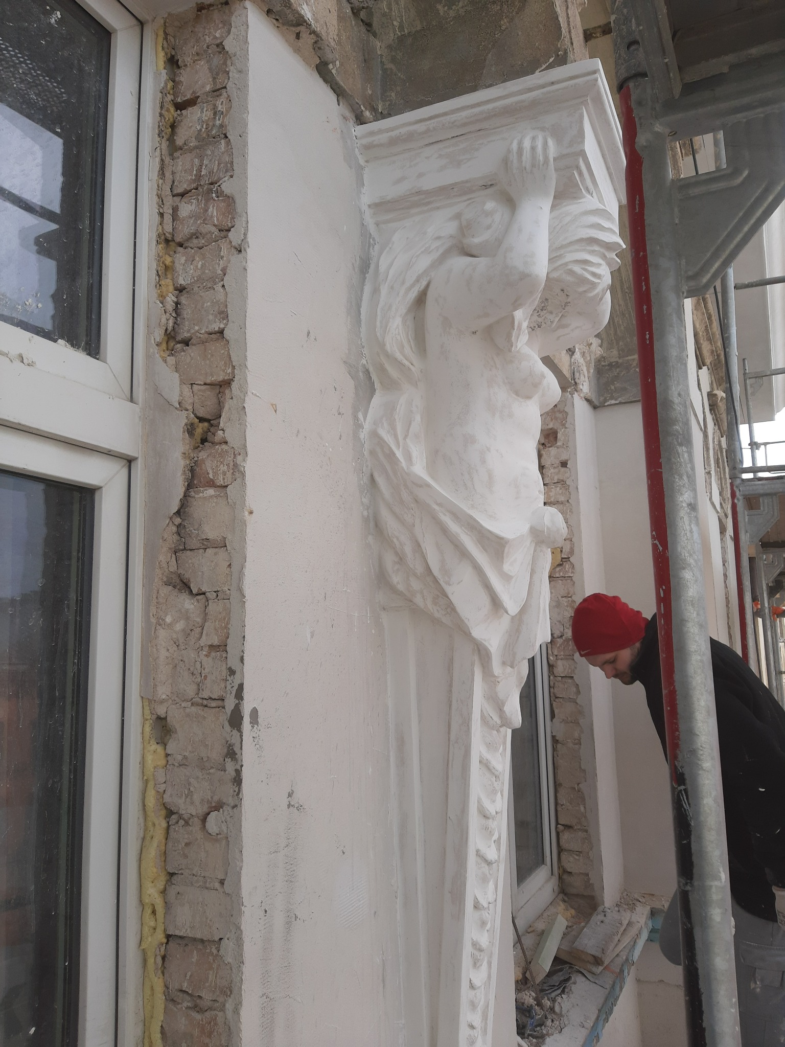 Renowacja elewacji kamienicy: pracownik w czerwonej czapce przy odnawianej kolumnie z postacią kobiecą, widoczne odsłonięte fragmenty muru i rusztowanie.