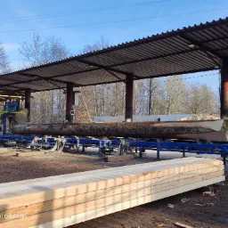 Proces obróbki drewna: kłoda na niebieskiej platformie w tartaku, obok stos tarcicy, w tle hałda trocin.