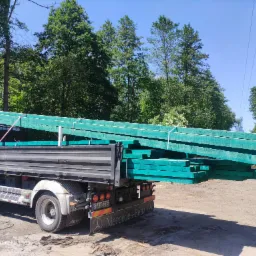 Ciężarówka Mercedes-Benz Atego z załadunkiem impregnowanej na zielono tarcicy budowlanej na tle drzew.