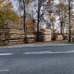 Stosy tarcicy ułożone wzdłuż drogi w jesiennej scenerii, z drzewami w tle i opadłymi liśćmi.