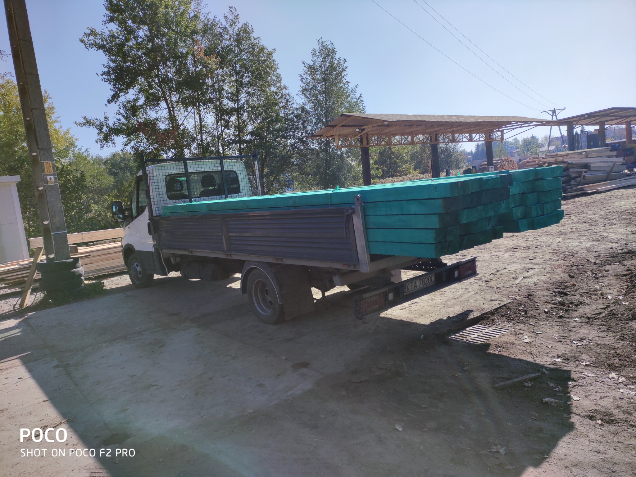 Ciężarówka z zieloną, impregnowaną tarcicą budowlaną na pace, zaparkowana na placu składowym z widocznymi belkami w tle.