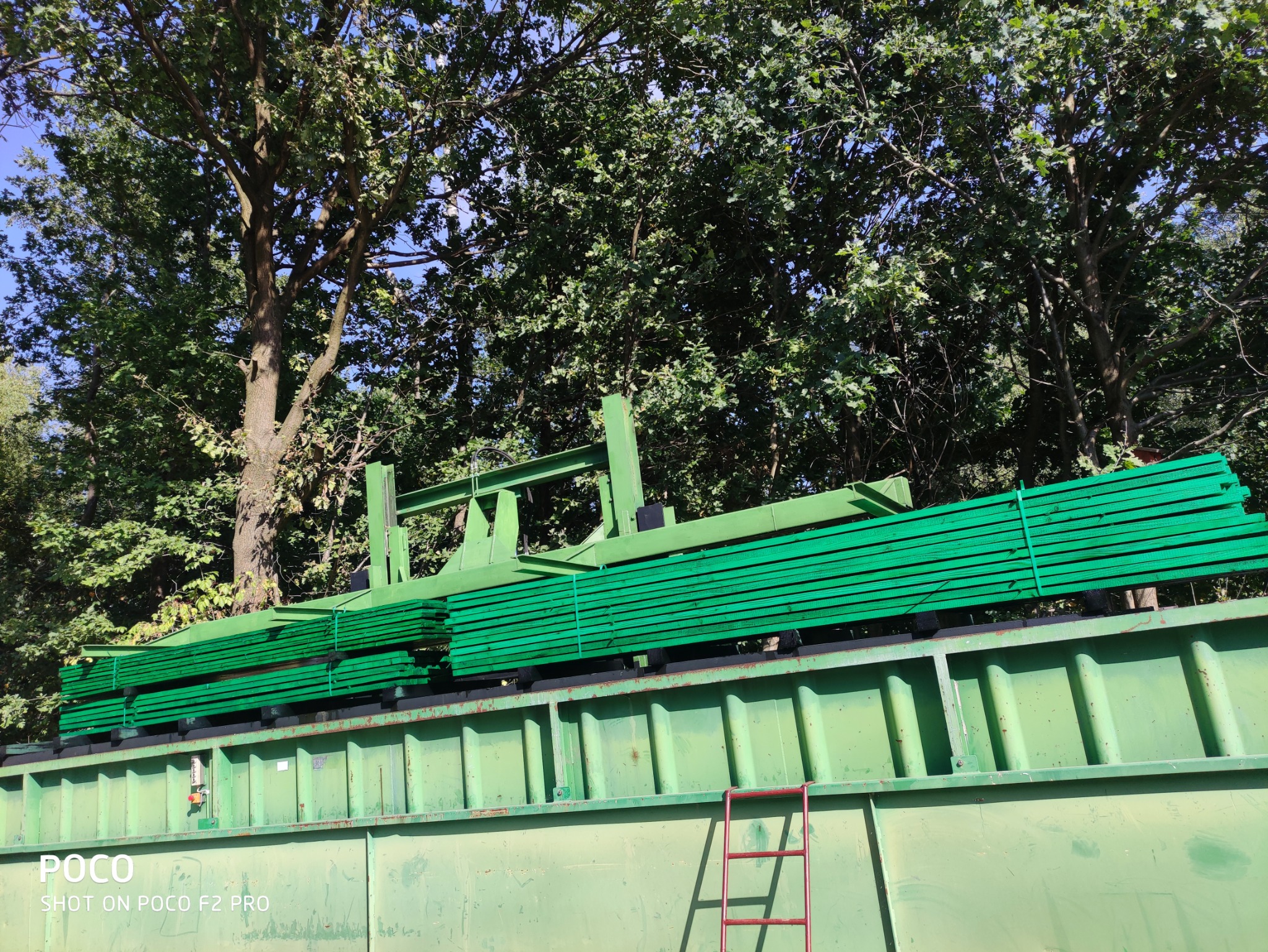 Załadunek zielonej, impregnowanej tarcicy na zieloną skrzynię transportową, widok z dołu na tle drzew.