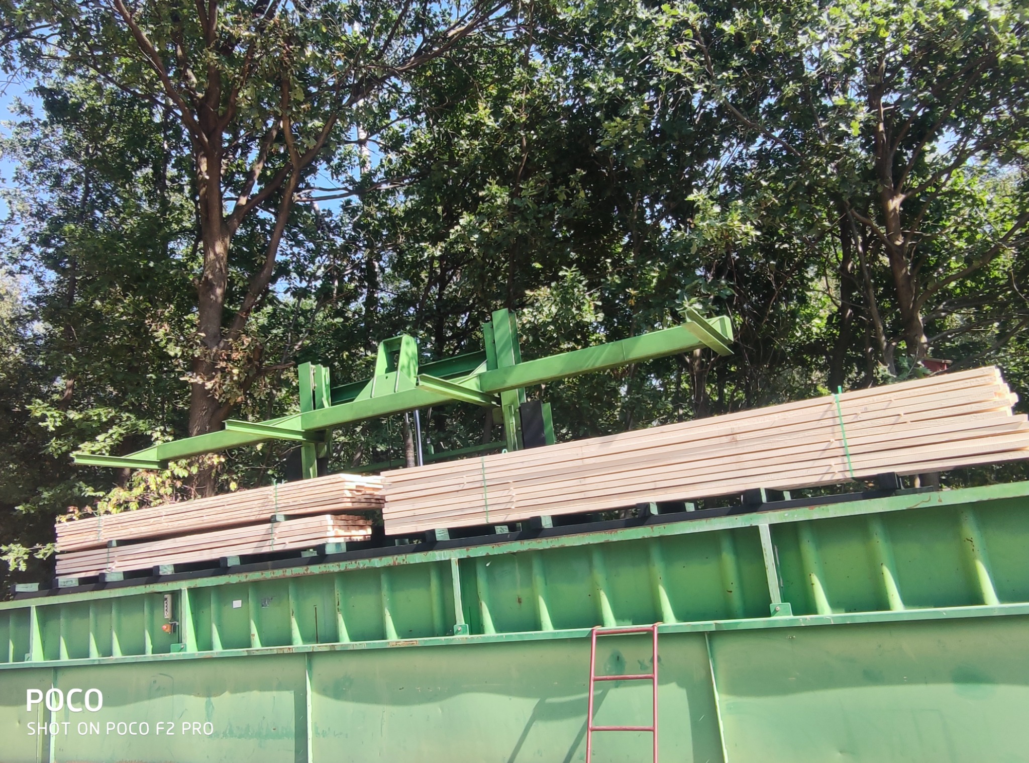 Stosy jasnej tarcicy na zielonym, metalowym pojeździe z dźwigiem, drzewa w tle.