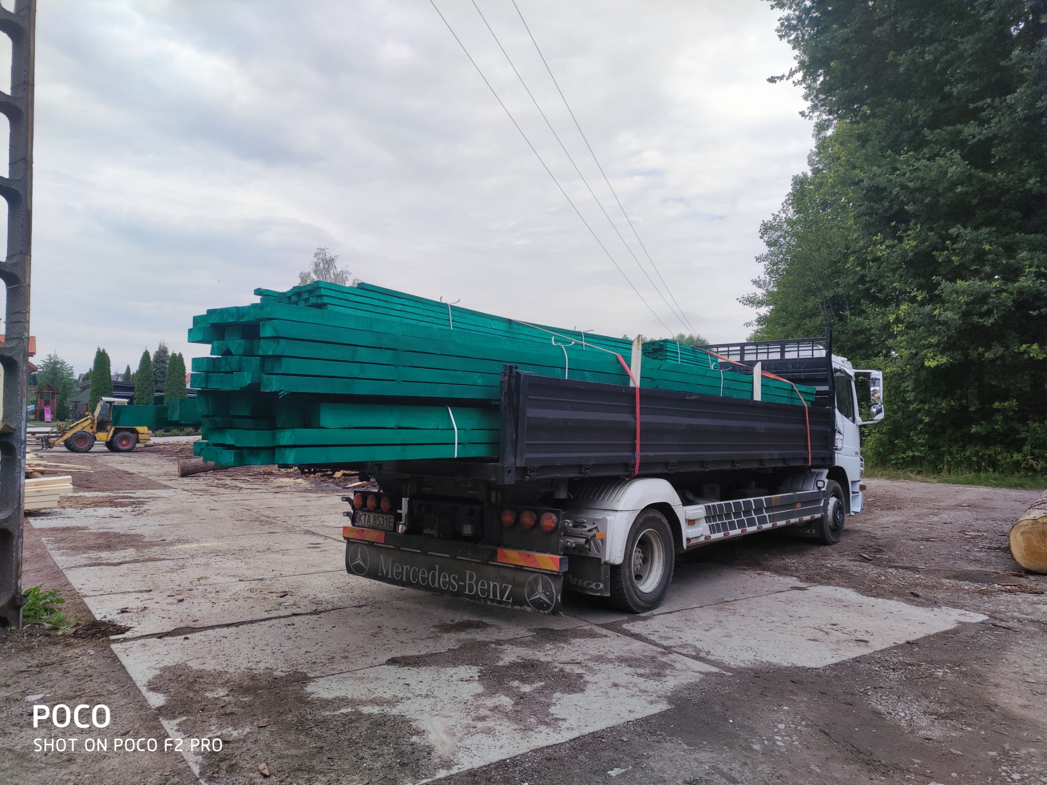Ciężarówka Mercedes-Benz Atego z załadunkiem impregnowanej na zielono tarcicy budowlanej, widok z tyłu na placu składowym.