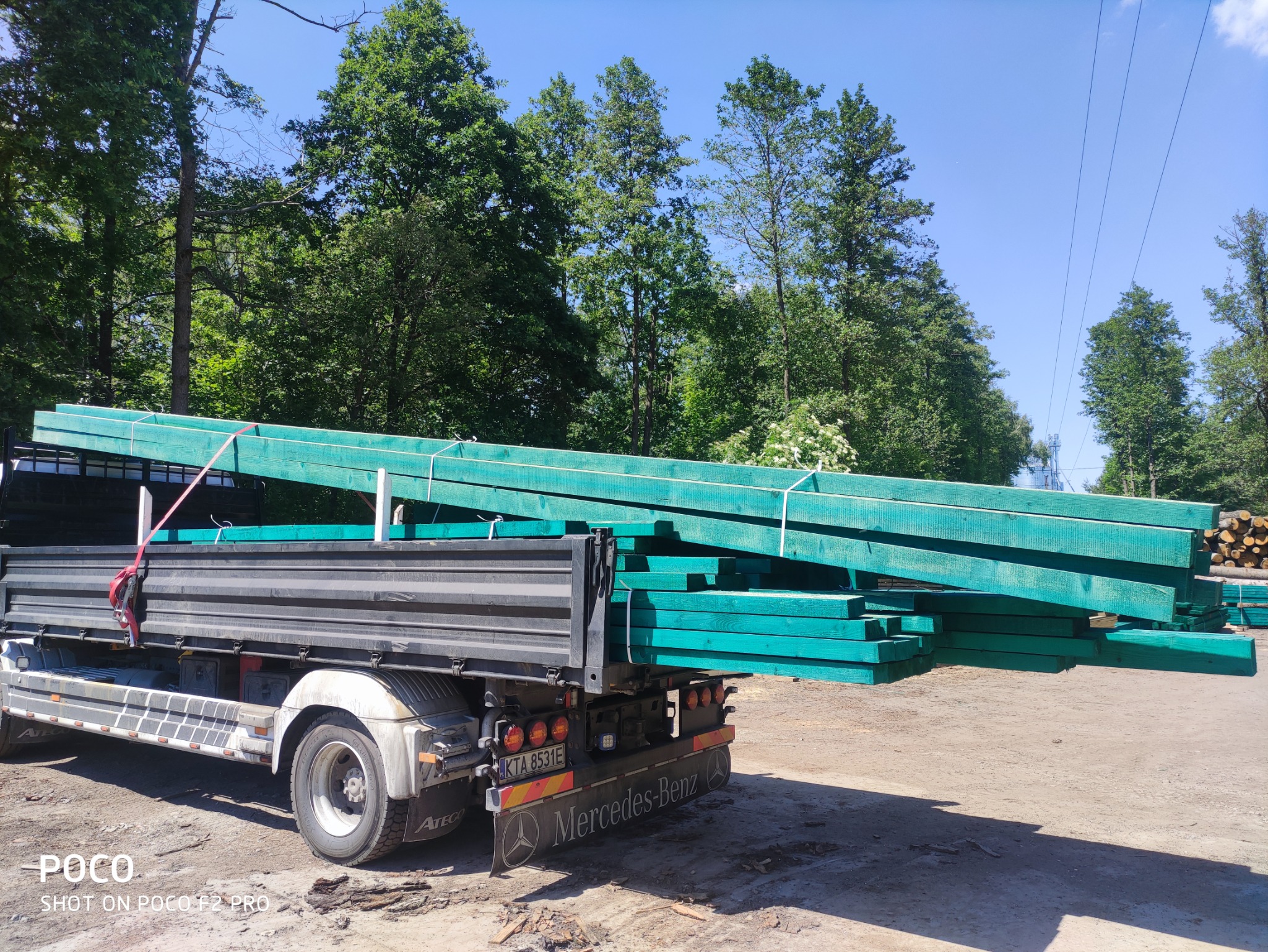 Ciężarówka Mercedes-Benz Atego z załadunkiem impregnowanej na zielono tarcicy budowlanej na tle drzew.