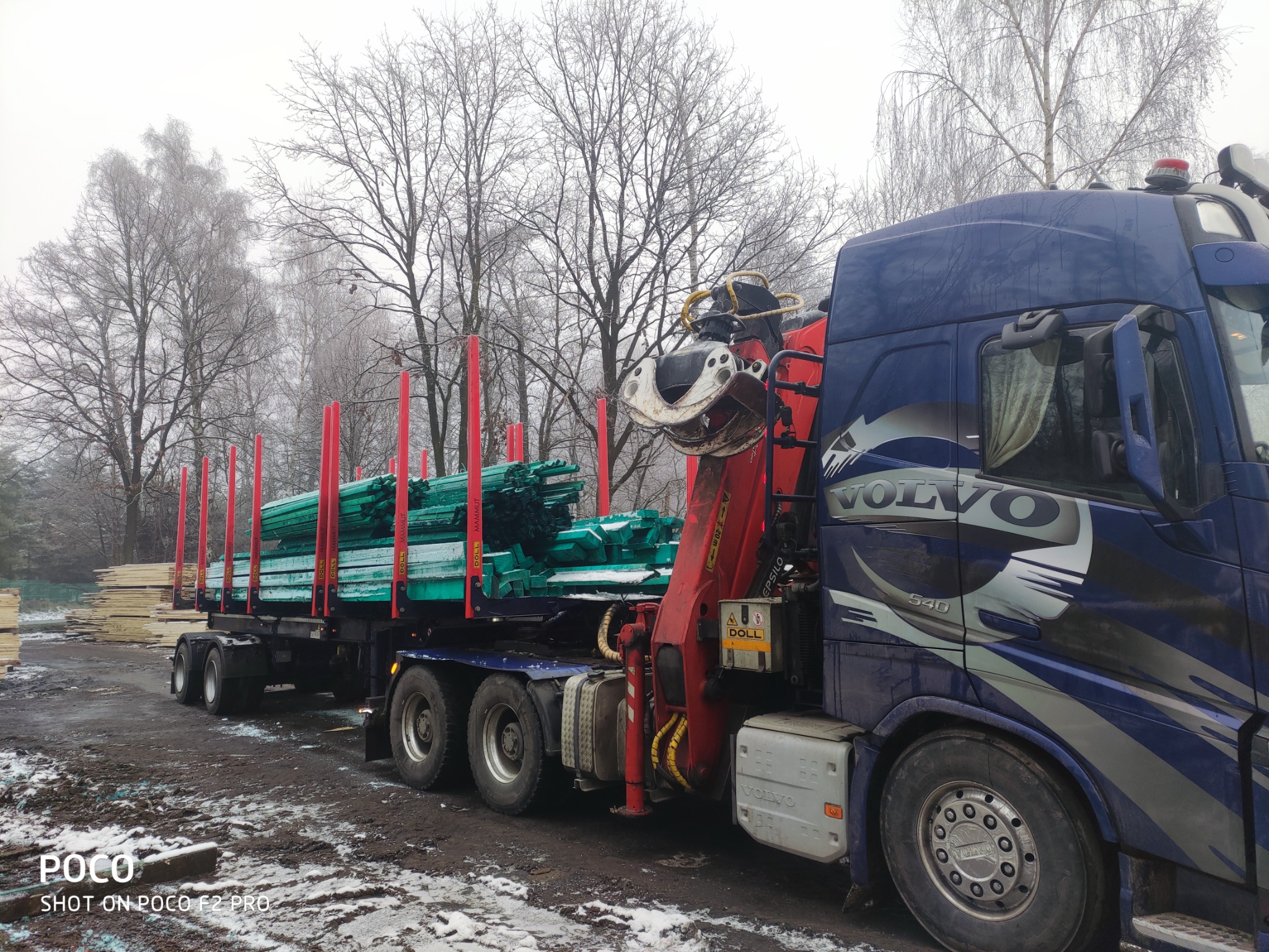Ciężarówka Volvo z hydraulicznym ramieniem załadowczym przewozi impregnowane, zielone belki na przyczepie w zimowy dzień.