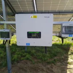 Biały inwerter Sofar Solar zamontowany na metalowej konstrukcji instalacji fotowoltaicznej, widoczny na tle paneli słonecznych i trawy, z dodatkowymi rozdzielnicami AC i DC marki Noark.