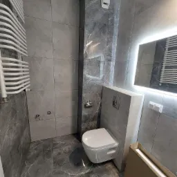 Nowoczesna łazienka wykończona szarymi płytkami imitującymi marmur, podwieszana toaleta, grzejnik drabinkowy i lustro z podświetleniem LED. Widoczny karton w rogu sugeruje świeżo zakończony montaż.