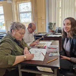 Trzy osoby siedzą przy stole, rysując schemat techniczny z użyciem szablonu i kolorowych kredek, w jasnym pomieszczeniu.