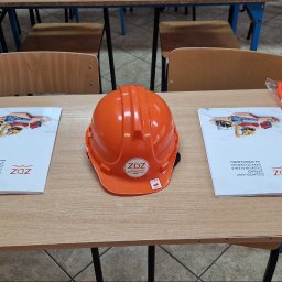 Dolnośląski Zakład Doskonalenia Zawodowego w Legnicy - Pomarańczowy kask budowlany z logo ZDZ, dwie pomarańczowe kamizelki odblaskowe w folii oraz dwie broszury ZDZ leżą na drewnianym biurku w sali szkoleniowej.