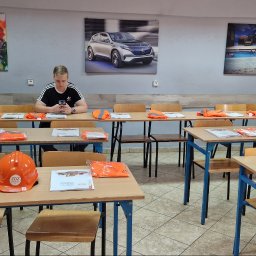 Dolnośląski Zakład Doskonalenia Zawodowego w Legnicy - Sala szkoleniowa z rzędami stołów, na których leżą pomarańczowe kamizelki odblaskowe i materiały szkoleniowe ZDZ; na pierwszym planie pomarańczowy kask; w tle siedzi mężczyzna w czarnej koszulce...