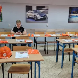 Sala szkoleniowa z rzędami stołów, na których leżą pomarańczowe kamizelki odblaskowe i materiały szkoleniowe ZDZ; na pierwszym planie pomarańczowy kask; w tle siedzi mężczyzna w czarnej koszulce...