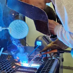 Spawacz w trakcie pracy, iskry i dym unoszą się znad metalu, ochronna osłona na szyi, przewód wentylacyjny nad głową