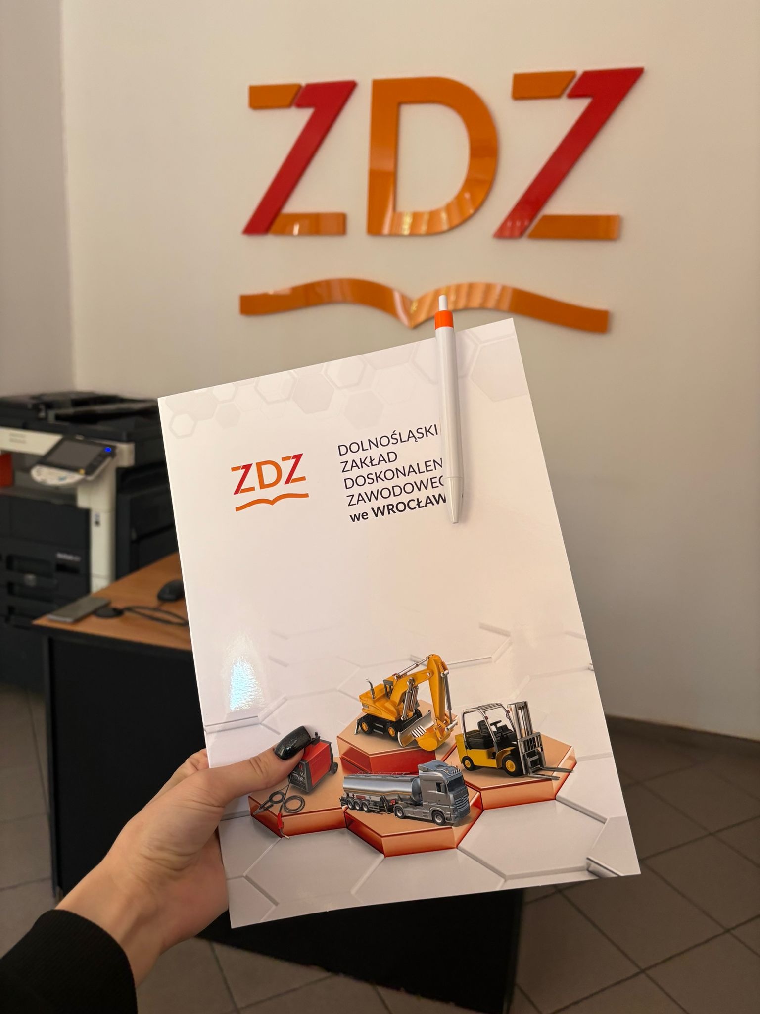 Osoba trzyma ulotkę Dolnośląskiego Zakładu Doskonalenia Zawodowego we Wrocławiu z grafikami maszyn budowlanych, na tle ściany z logo ZDZ i drukarki biurowej.