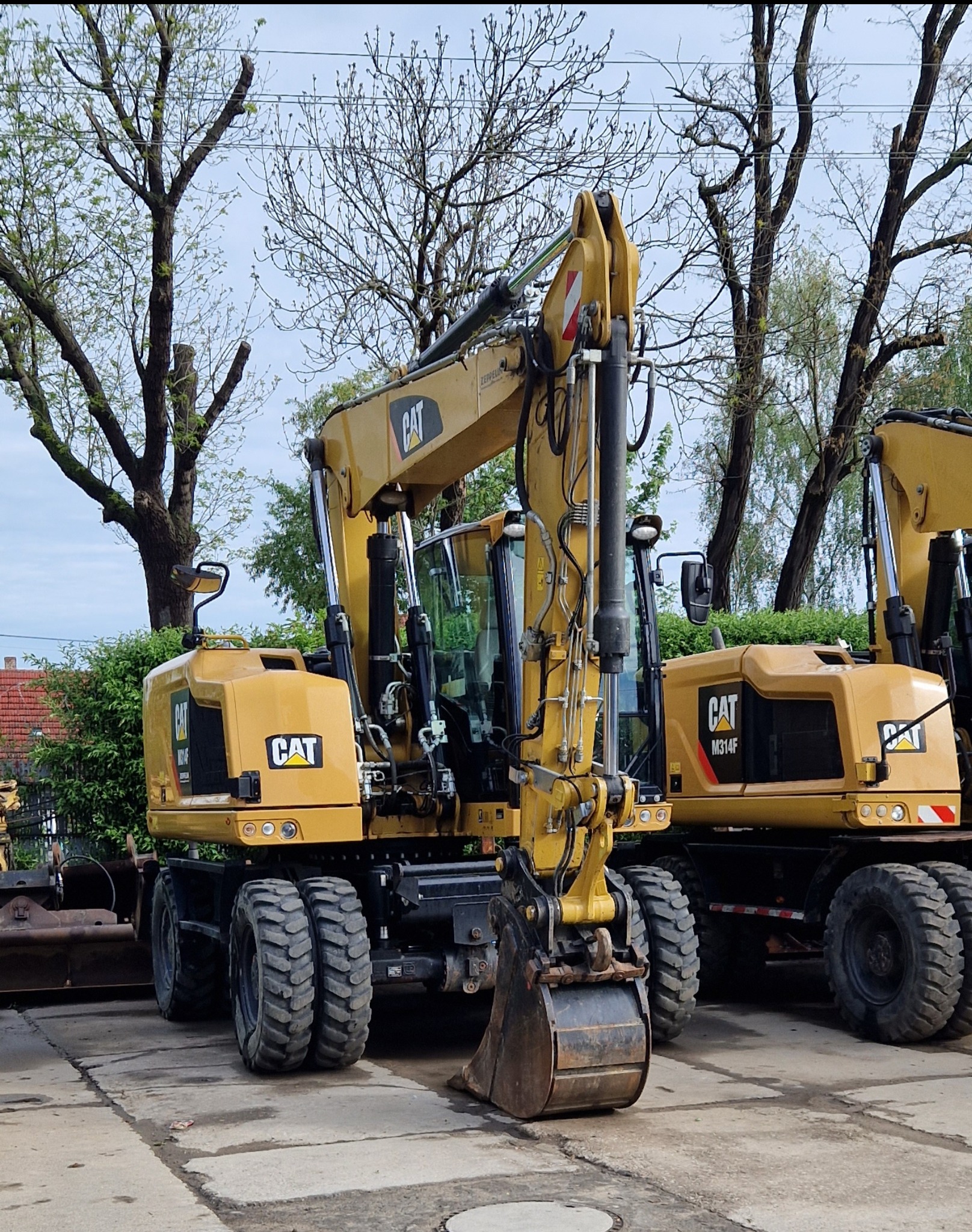 Żółta koparka kołowa CAT M314F zaparkowana na betonowej nawierzchni, widoczne drzewa w tle.
