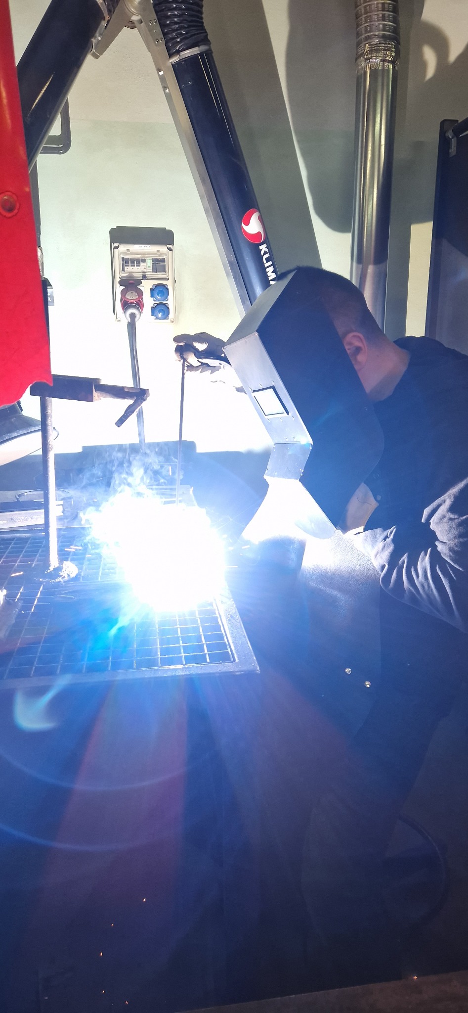 Spawacz w masce ochronnej spawa metalową konstrukcję na kratownicy, widoczny system odciągu spalin KLiMA, iskry i jasne światło spawania.