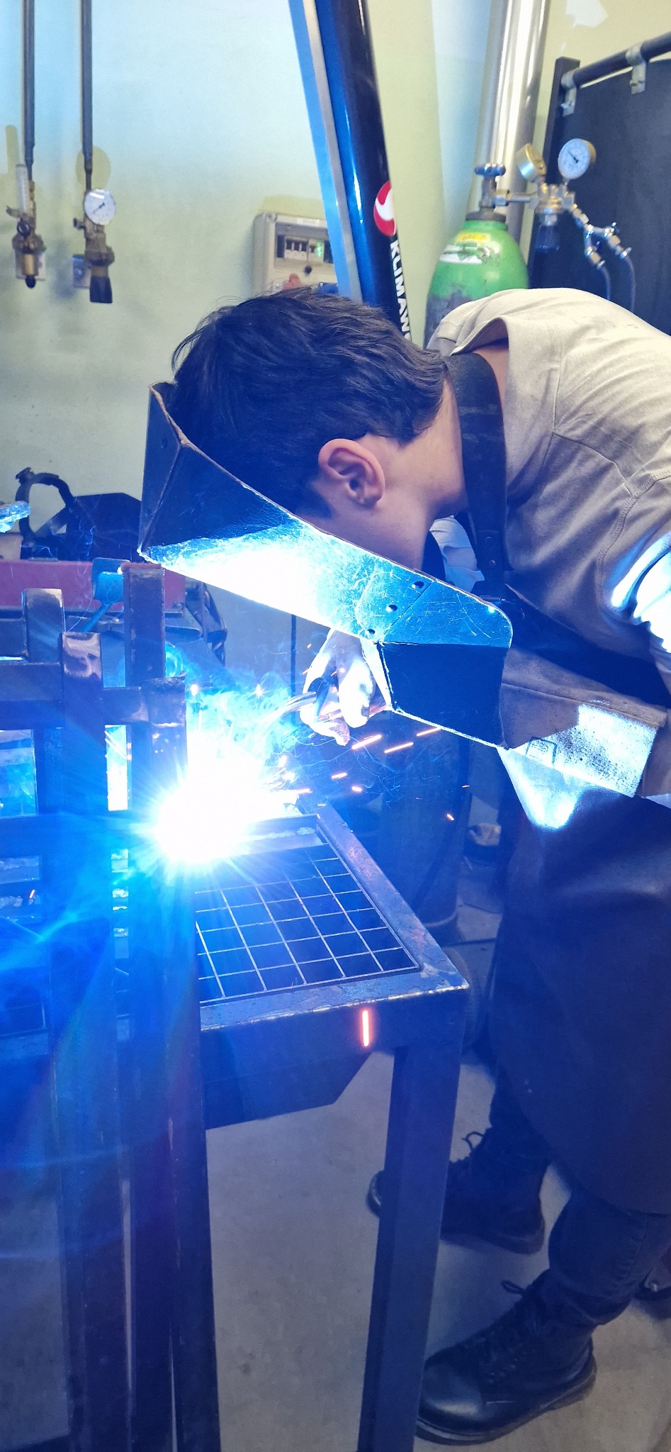 Spawacz w masce spawalniczej pochylony nad metalową konstrukcją, widoczne iskry i intensywne światło spawania, butla z gazem w tle.