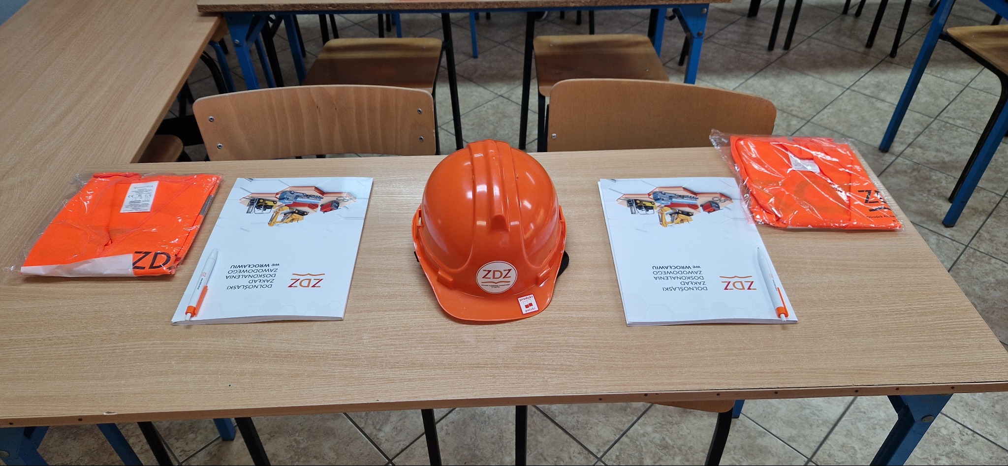 Pomarańczowy kask budowlany z logo ZDZ, dwie pomarańczowe kamizelki odblaskowe w folii oraz dwie broszury ZDZ leżą na drewnianym biurku w sali szkoleniowej.
