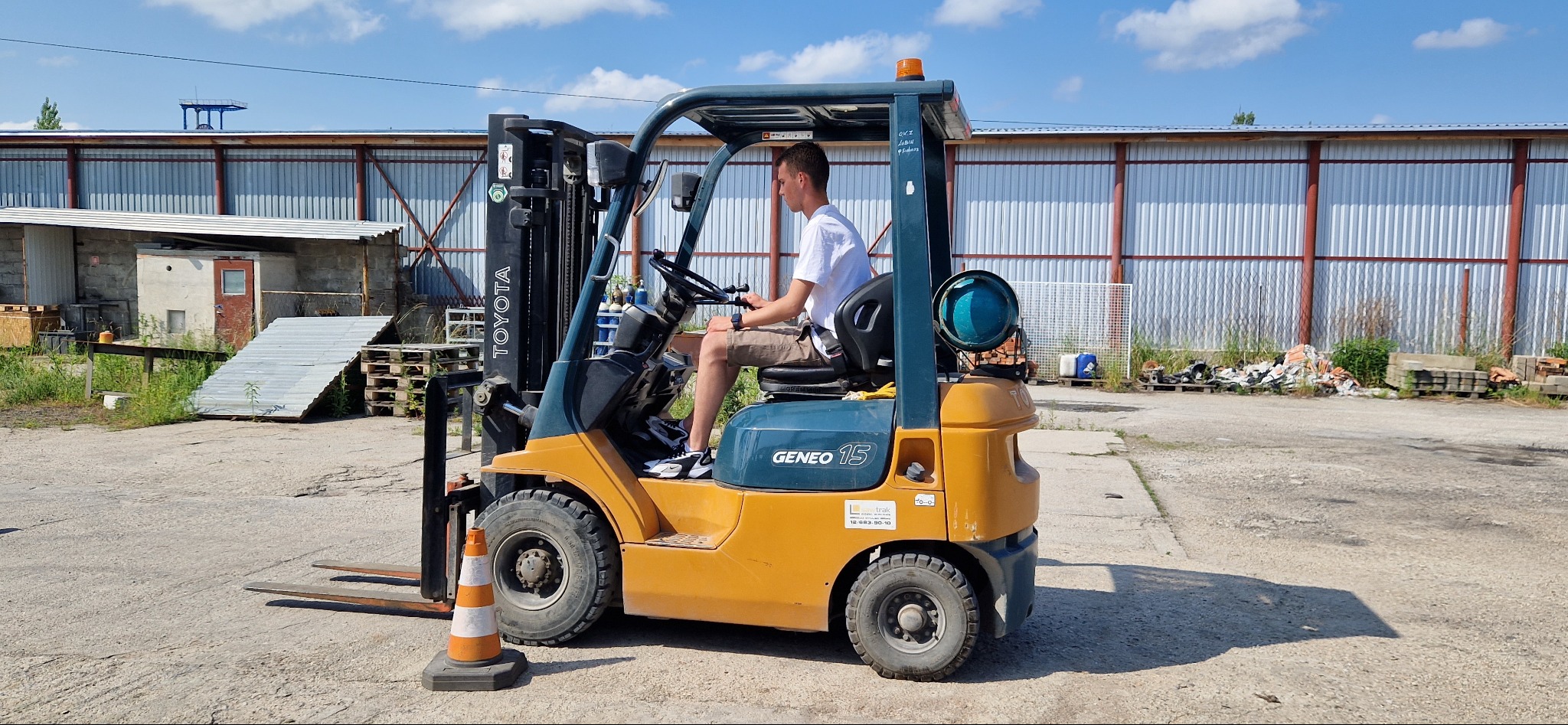 Młody mężczyzna w białej koszulce i beżowych spodenkach obsługuje wózek widłowy Toyota GENEO 15 na placu budowy, obok stoi pomarańczowy pachołek, w tle hala z blachy falistej.