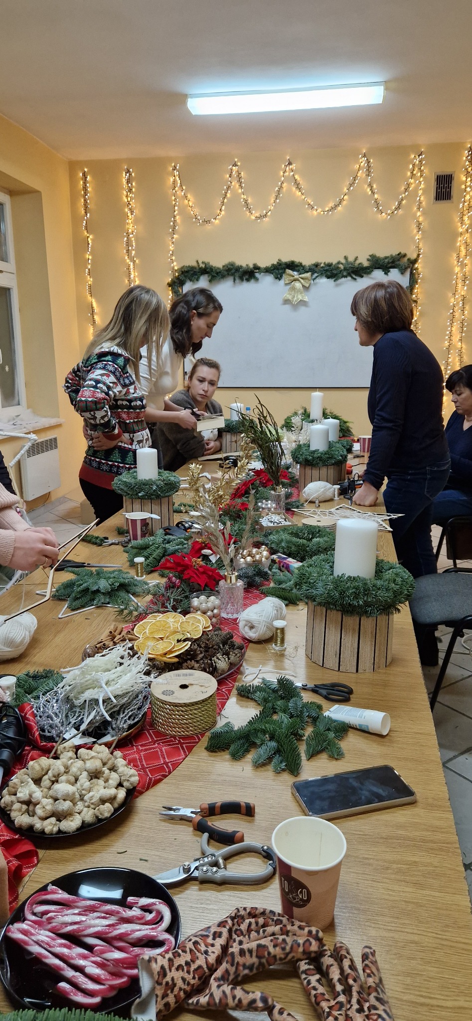 Grupa osób uczestniczy w warsztatach rękodzielniczych, tworząc dekoracje świąteczne ze świerkowych gałązek, suszonych pomarańczy, szyszek i świec na drewnianym stole ozdobionym świetlnymi girlandami.