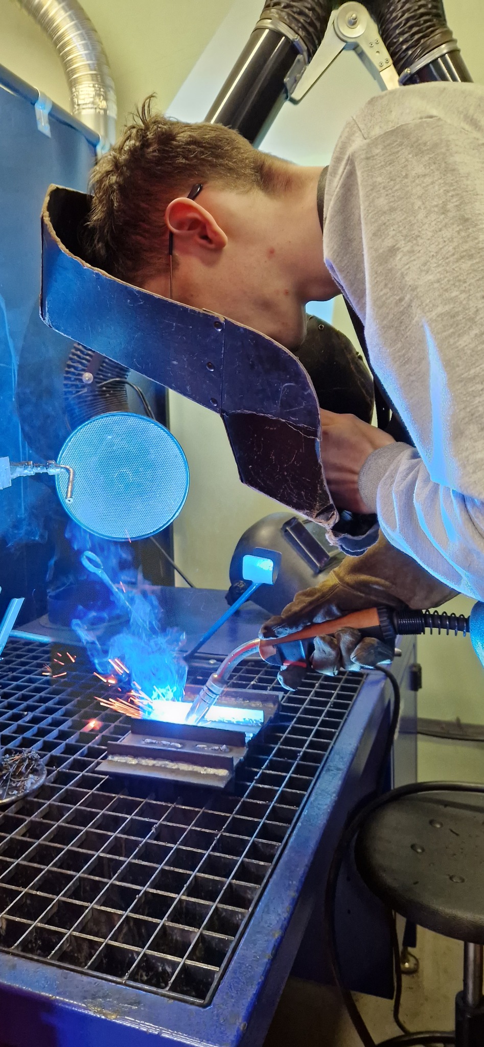 Spawacz w trakcie pracy, iskry i dym unoszą się znad metalu, ochronna osłona na szyi, przewód wentylacyjny nad głową