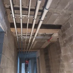 Równoległe rzędy rur instalacji wodnej i grzewczej zamocowane do sufitu w surowym wnętrzu, widoczny fragment instalatora na drabinie w oddali.