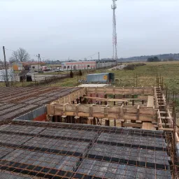Zbrojenie stropu w szalunku, widoczne bloczki betonowe i stalowe pręty, tło stanowią budynki gospodarcze i maszt telekomunikacyjny w oddali.