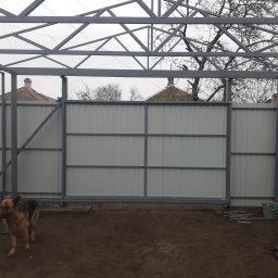 Progres Vitalii Morozov - W trakcie montażu: szara, metalowa konstrukcja bramy przesuwnej z wypełnieniem z blachy trapezowej, widoczny pies owczarek niemiecki na tle ziemi i elementów budowlanych.