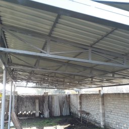 Progres Vitalii Morozov - Konstrukcja zadaszenia z metalowych profili i blachy trapezowej, widok od dołu, pracownik na drabinie w tle, murowany płot w otoczeniu.