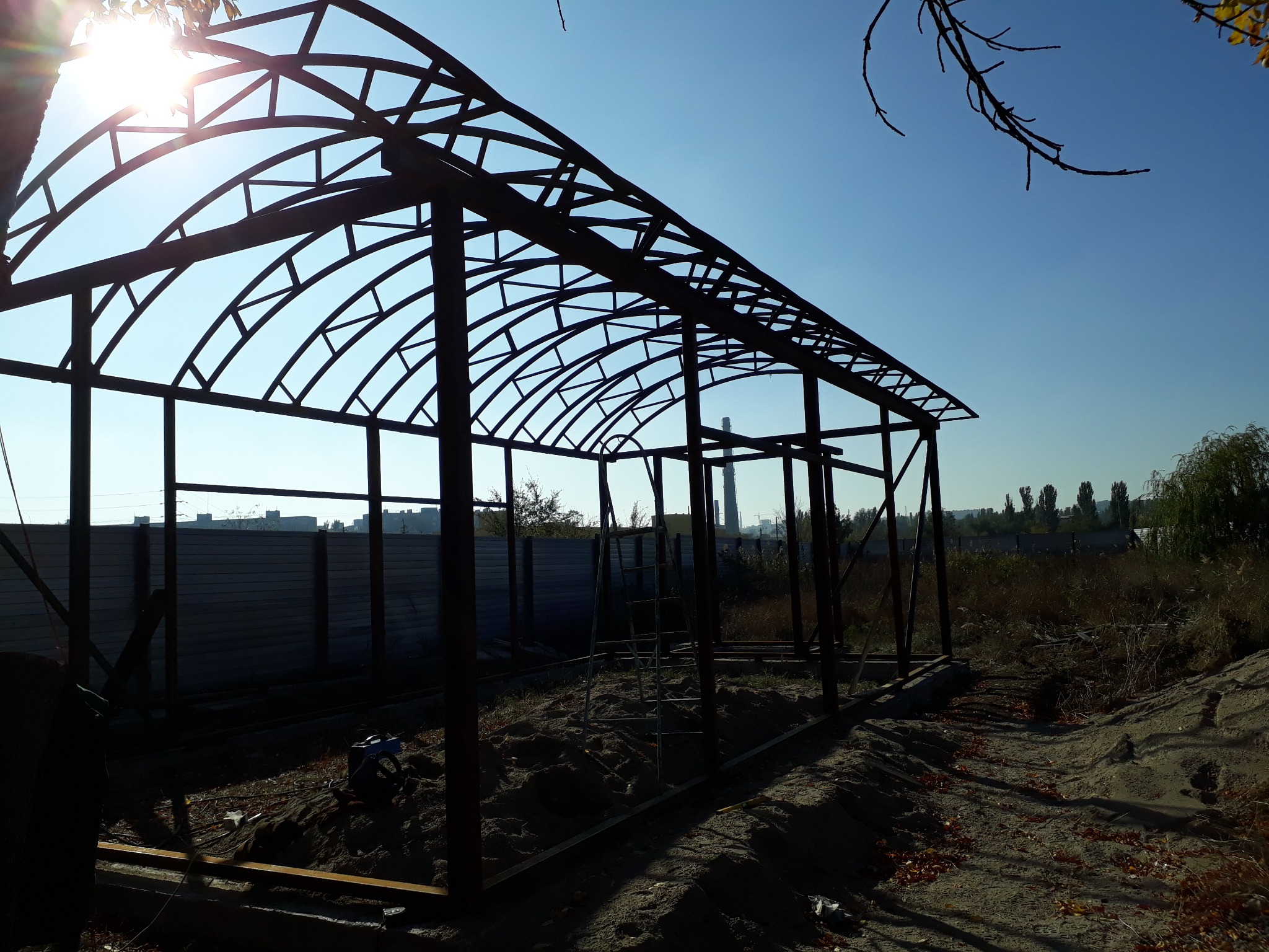 Konstrukcja metalowa z łukowatym dachem w trakcie budowy, widoczna drabina, w tle betonowe ogrodzenie i zabudowania miejskie w oddali, jasne słońce.