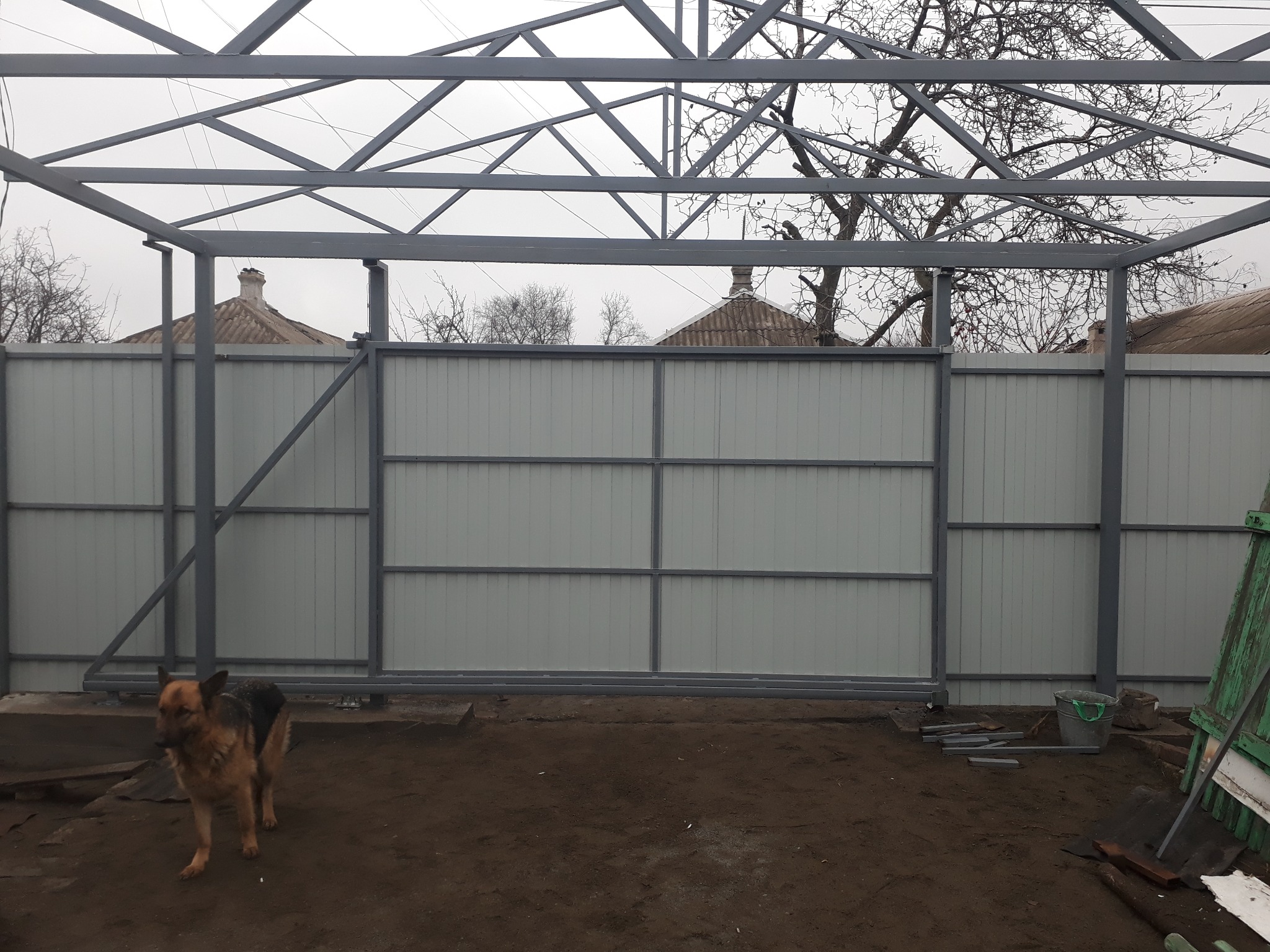 W trakcie montażu: szara, metalowa konstrukcja bramy przesuwnej z wypełnieniem z blachy trapezowej, widoczny pies owczarek niemiecki na tle ziemi i elementów budowlanych.