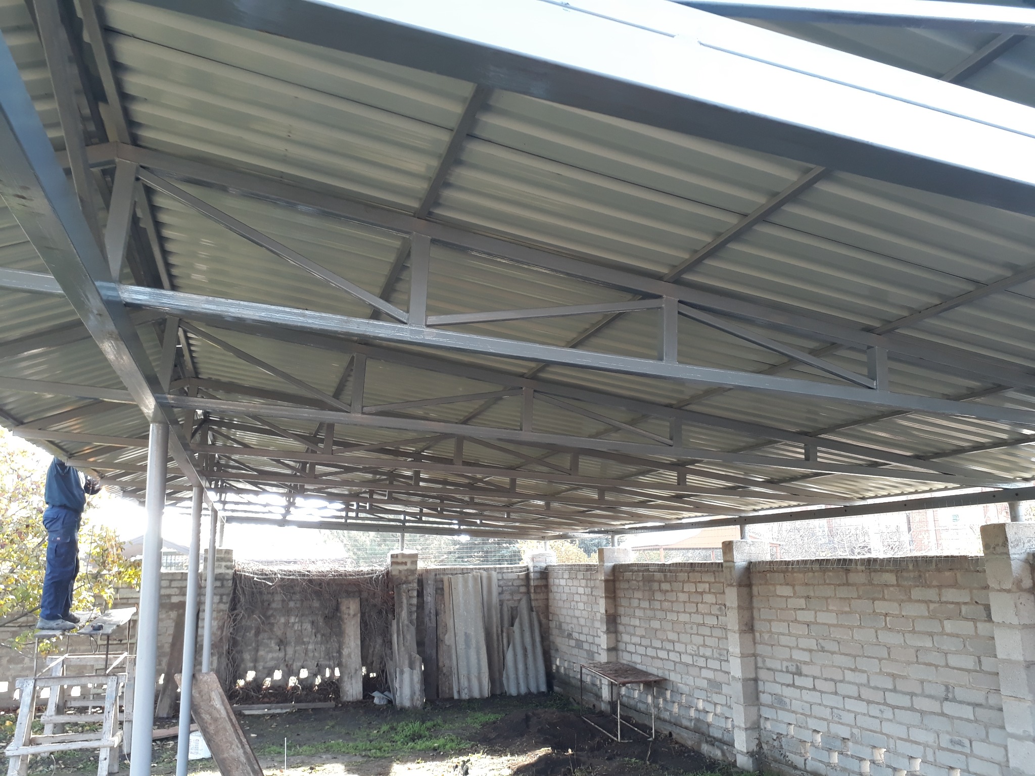 Konstrukcja zadaszenia z metalowych profili i blachy trapezowej, widok od dołu, pracownik na drabinie w tle, murowany płot w otoczeniu.