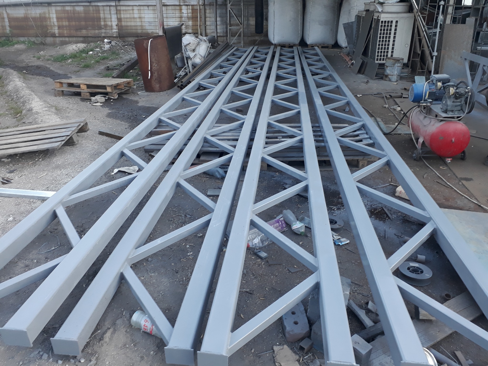 Sześć pomalowanych na szaro, stalowych, kratownicowych elementów konstrukcyjnych leży na ziemi w hali produkcyjnej. W tle widoczne białe worki, palety i czerwony kompresor.