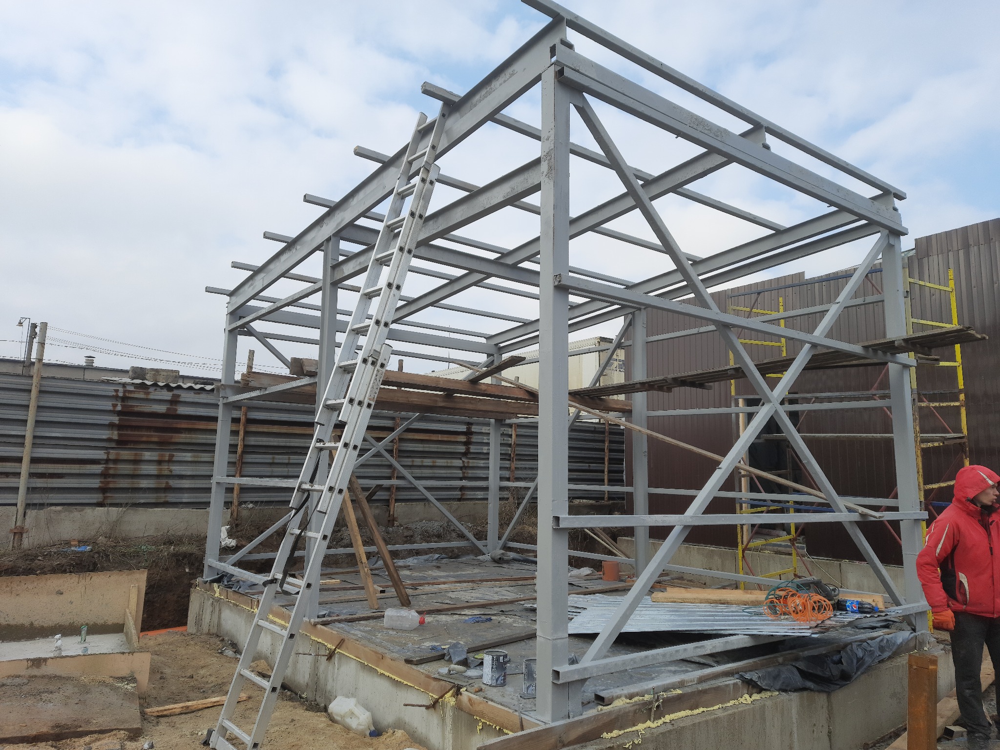 Konstrukcja szkieletowa z profili stalowych w trakcie montażu, widoczna drabina oparta o konstrukcję, teren budowy z widocznym wykopem pod fundamenty i częściowo ułożoną podłogą.