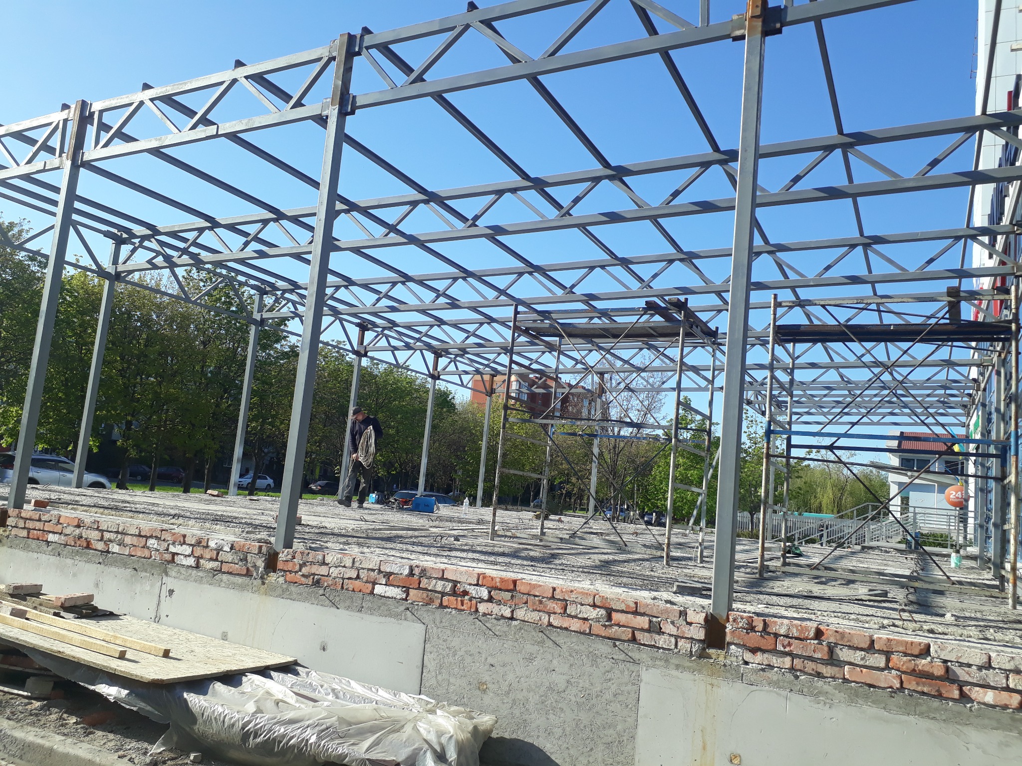 Konstrukcja stalowa budynku w trakcie budowy, widoczne rusztowania, ceglana podmurówka i pracownik z liną na tle zieleni i błękitnego nieba.