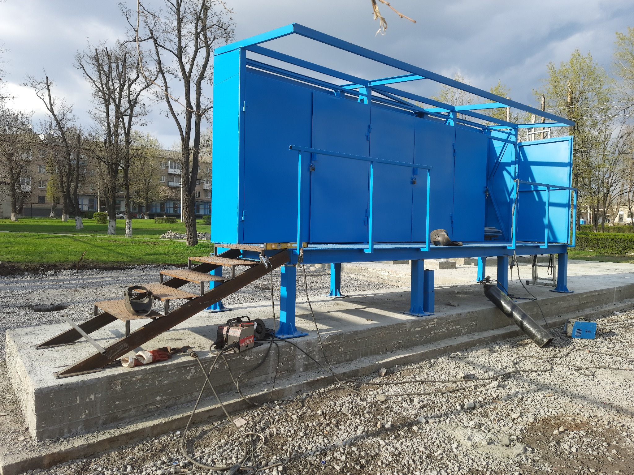 Niebieska metalowa konstrukcja z drzwiami i schodami na betonowym fundamencie, w trakcie montażu, widoczny sprzęt spawalniczy i butla gazowa, w tle drzewa i budynek mieszkalny.