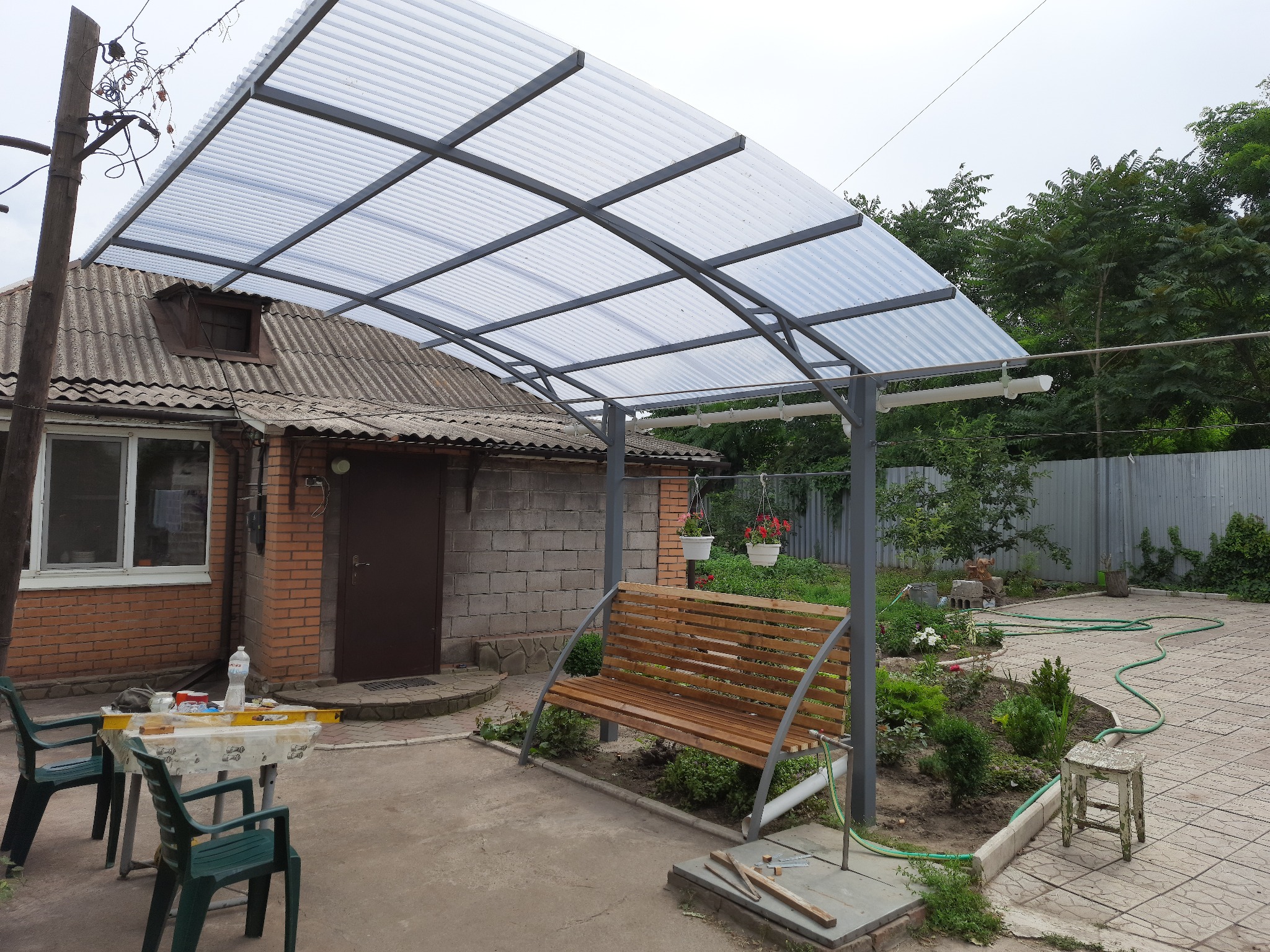 Zadaszenie tarasu z poliwęglanu falistego na metalowej konstrukcji, widoczny drewniany stół z krzesłami, ogród i dom z cegły w tle.