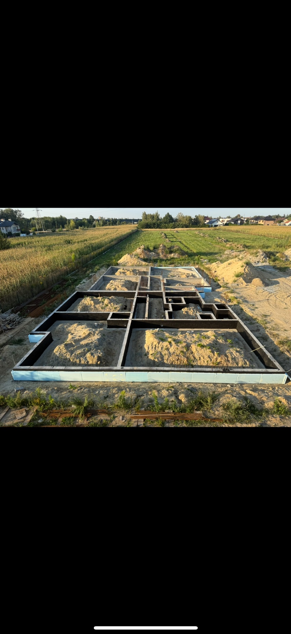 Zalane fundamenty domu jednorodzinnego na piaszczystym terenie, widok z góry. Gotowy szalunek z izolacją, wypełniony piaskiem. W tle łąka i zabudowania.