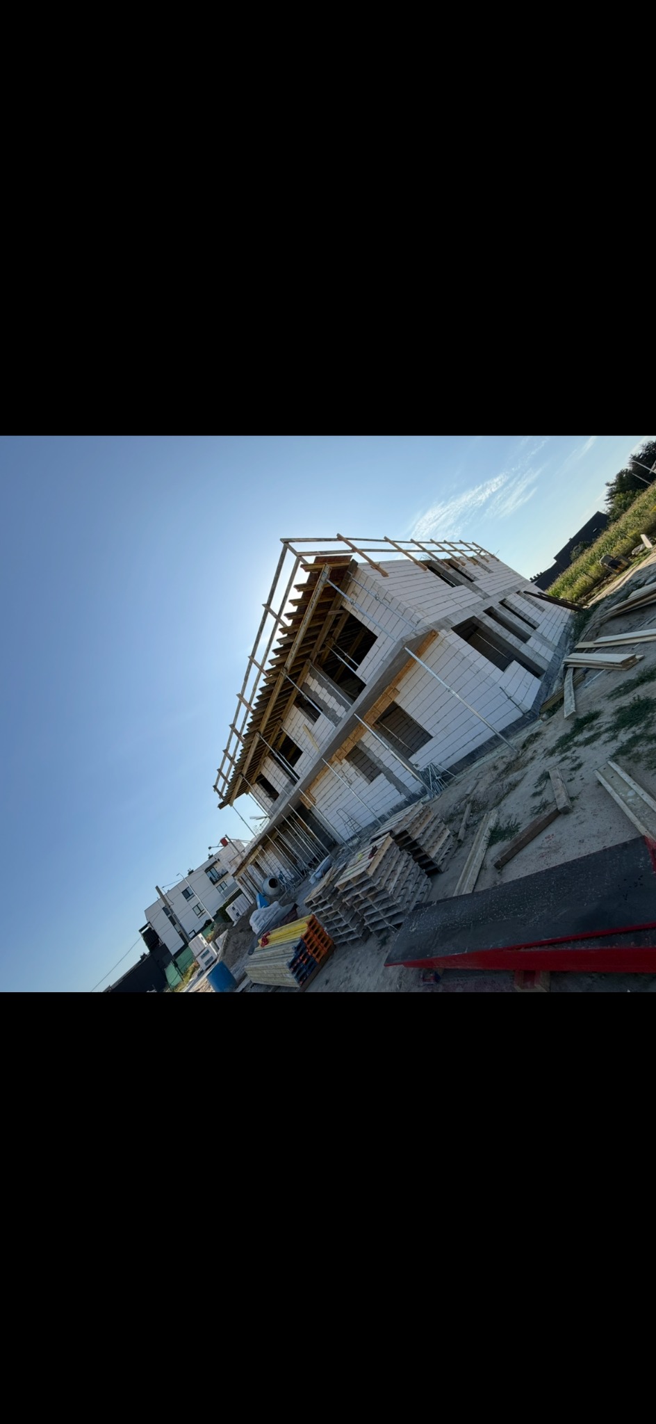 Wznoszenie dwukondygnacyjnego domu z białych bloczków, widok z dołu, niebo w tle. Częściowo zrobiona więźba dachowa. Materiały budowlane obok.