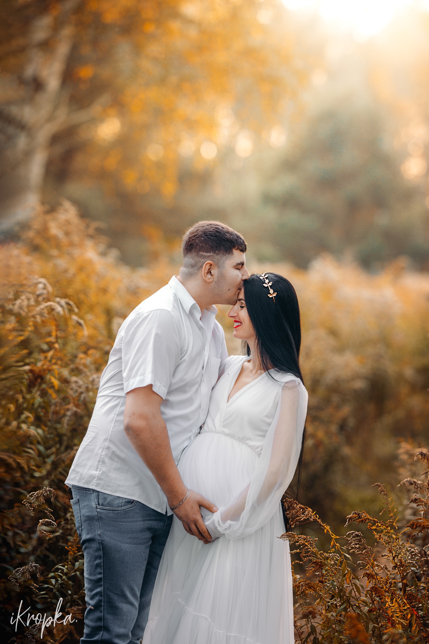 Przyszły tata całuje w czoło uśmiechniętą, elegancką ciężarną w białej sukni na tle jesiennego krajobrazu.