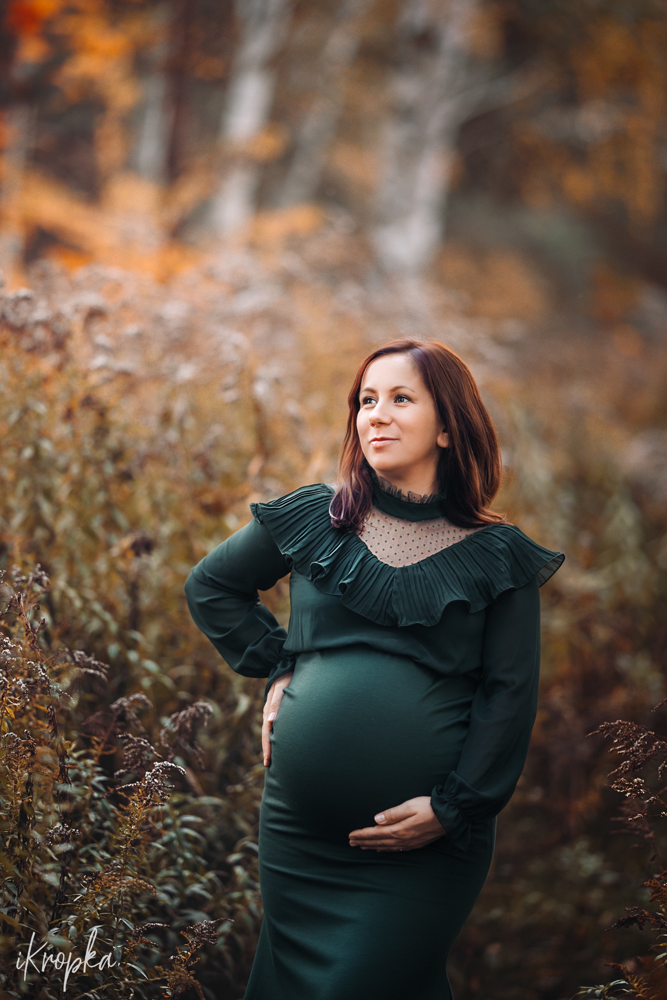 Portret kobiety w ciąży w zielonej sukience z falbanami, pozującej na tle jesiennego krajobrazu z drzewami i krzewami w odcieniach brązu i pomarańczy.