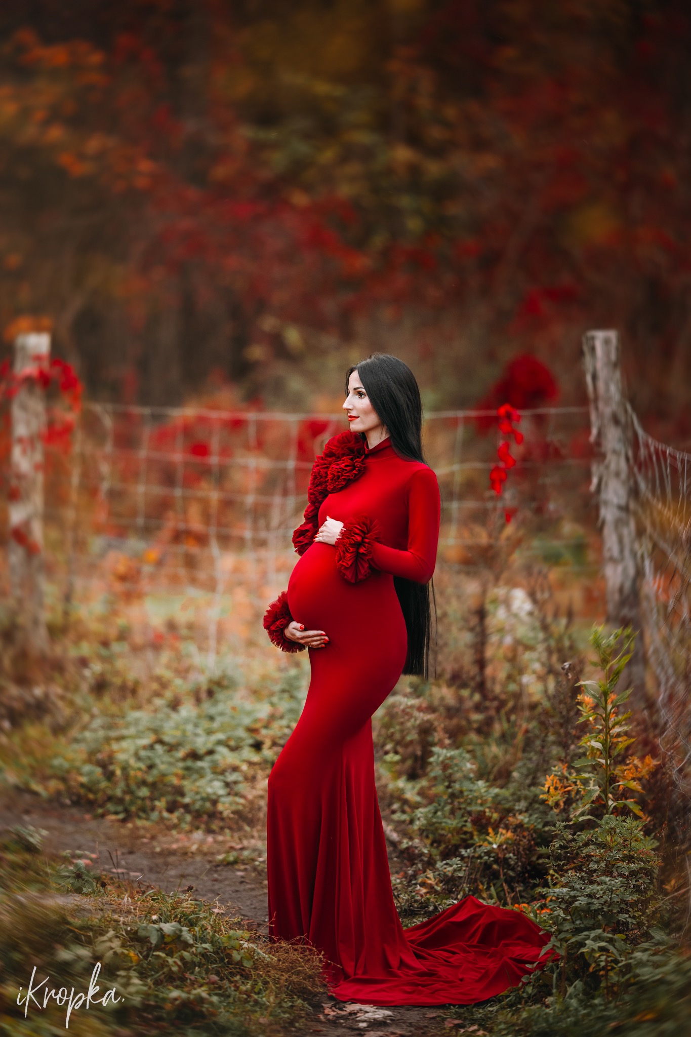 Elegancka kobieta w czerwonej sukni ciążowej pozuje na tle jesiennego krajobrazu z drewnianym płotem i czerwonymi liśćmi.