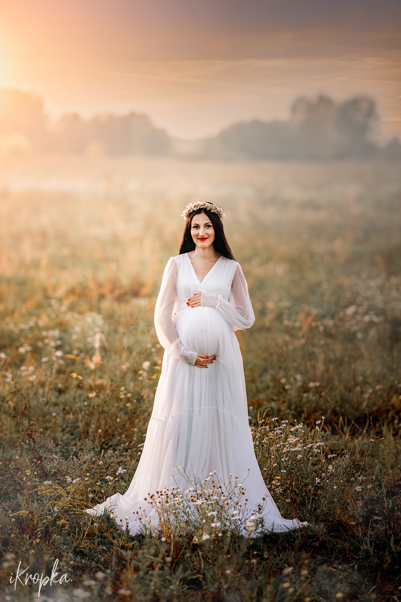 Uśmiechnięta kobieta w białej sukni ciążowej z wiankiem na głowie, stojąca na łące w promieniach zachodzącego słońca.