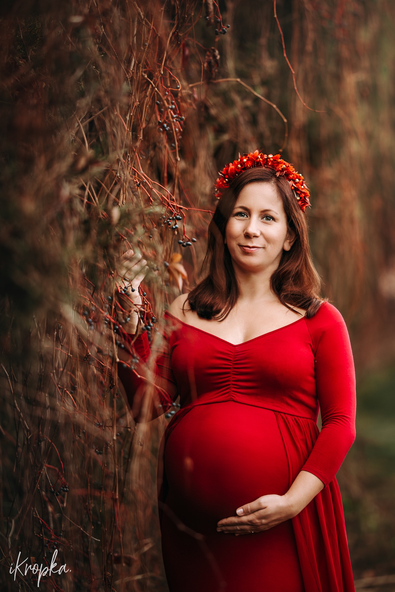 Uśmiechnięta kobieta w czerwonej sukni ciążowej i wianku z czerwonych kwiatów, pozuje w plenerze przy gałęziach z owocami.