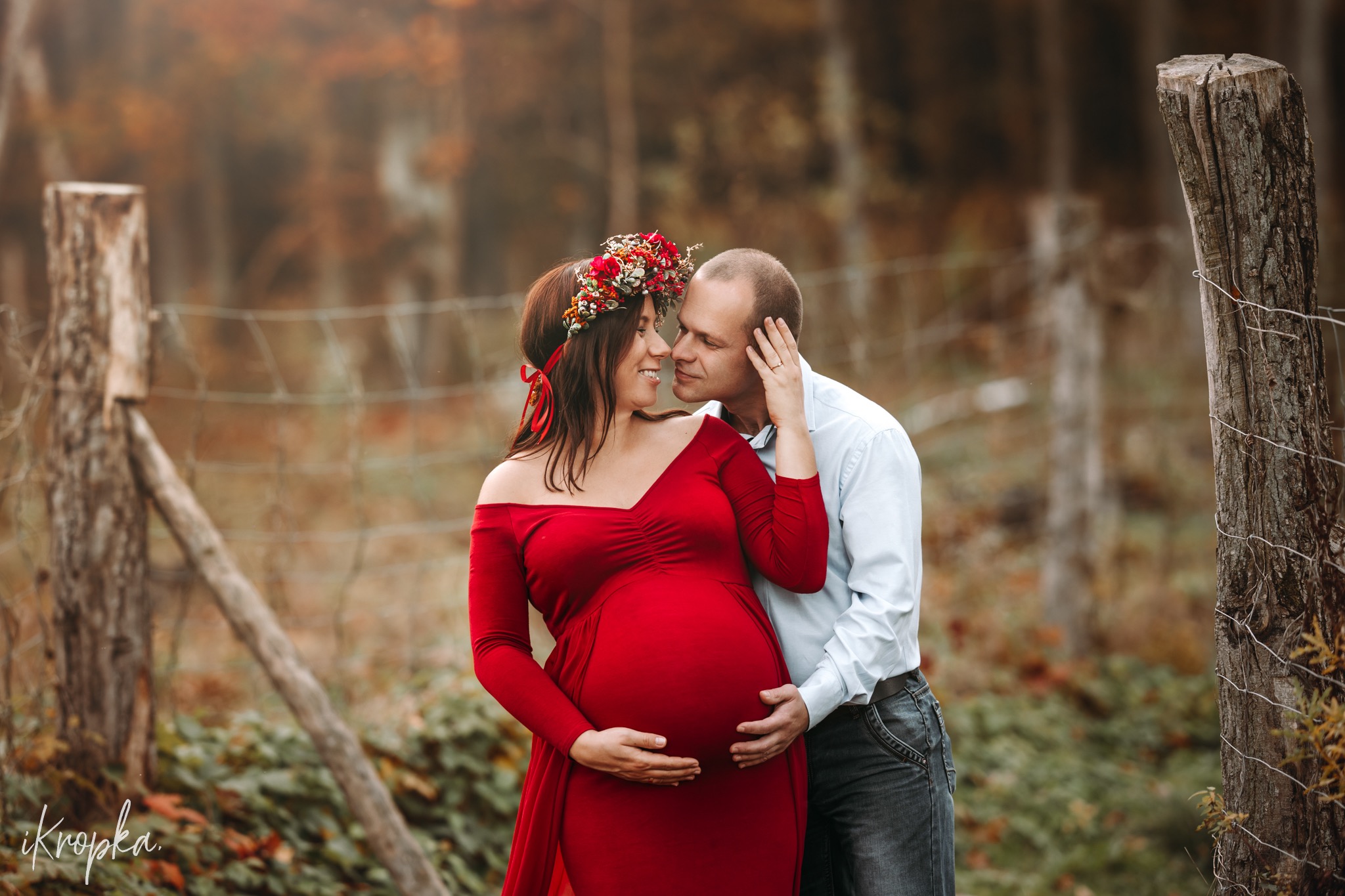 Kobieta w czerwonej sukni ciążowej i wianku z czerwonych kwiatów, wtulona w mężczyznę na tle jesiennego krajobrazu i drewnianego płotu.