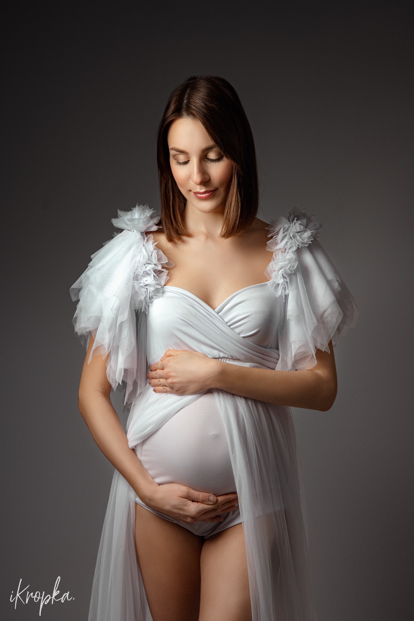 Kobieta w zwiewnej białej sukni z tiulowymi zdobieniami na ramionach, delikatnie obejmująca ciążowy brzuszek na szarym tle.