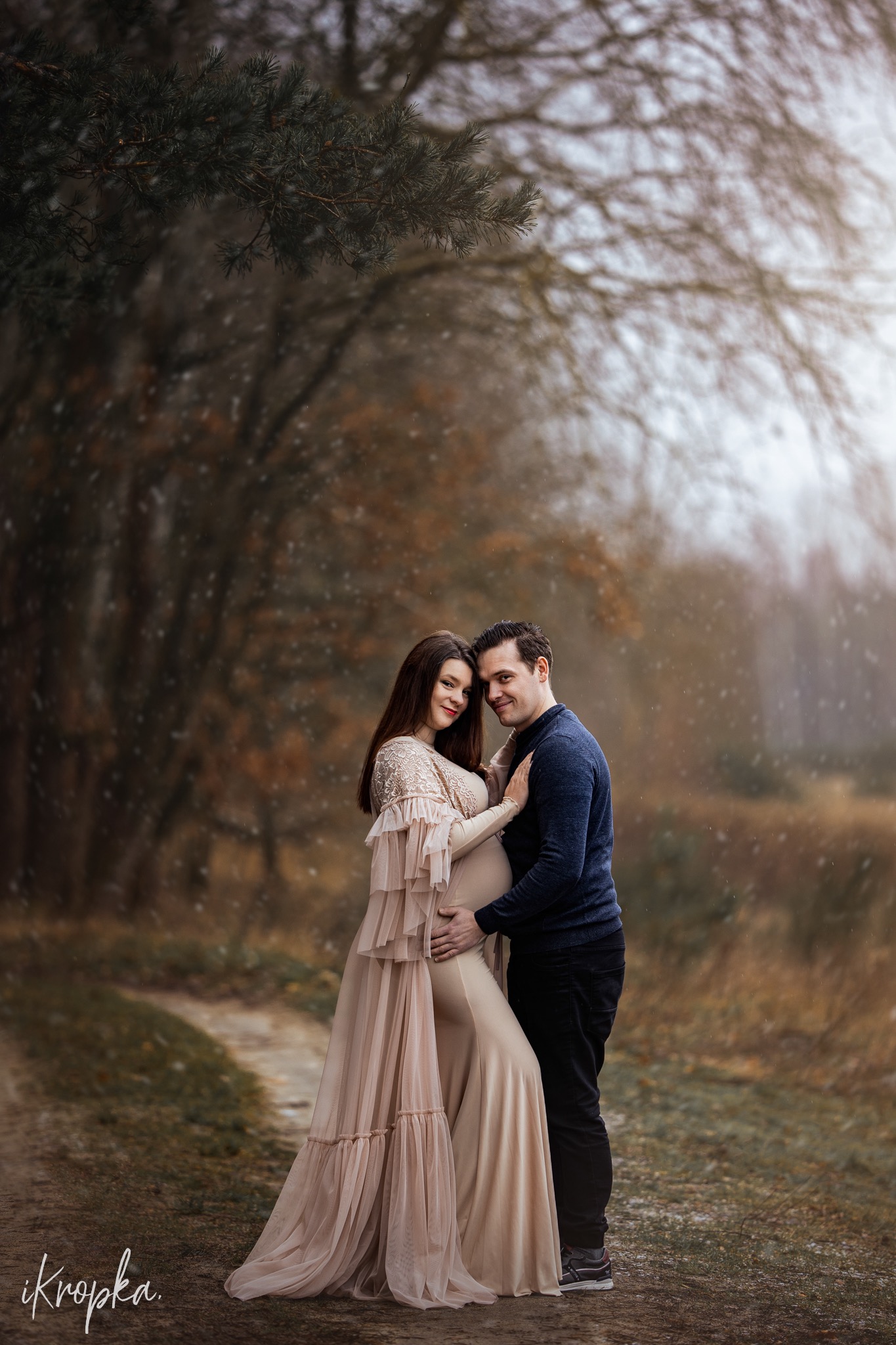 Ciążowa sesja plenerowa: przyszła mama w beżowej sukni z falbanami i koronkowym karczkiem pozuje z partnerem na leśnej ścieżce podczas padającego śniegu.
