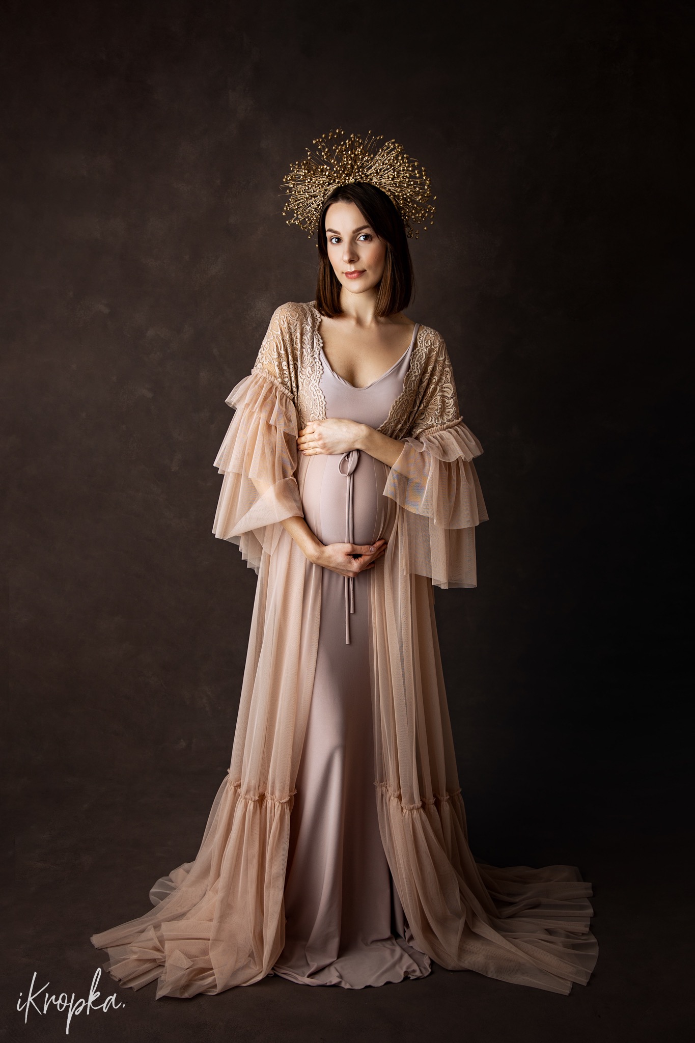 Portret kobiety w ciąży, w długiej sukni i koronkowym szlafroku, z ozdobną złotą koroną na głowie, na ciemnym, teksturowanym tle.