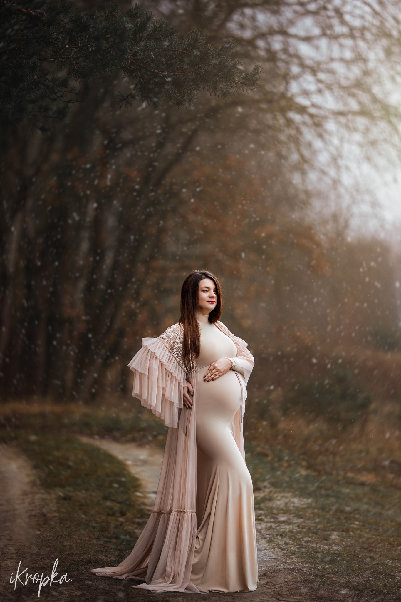 Kobieta w ciąży w długiej, beżowej sukni i narzutce z falbanami, pozuje na leśnej ścieżce podczas delikatnych opadów śniegu.